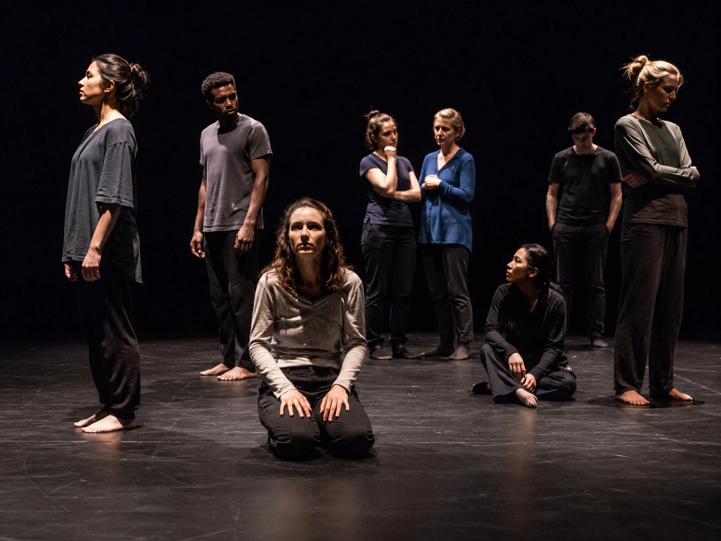 A live-theatre bare-stage concept for Find Me, with an adult ensemble spaced across an open black-box stage under practical theatre lighting.