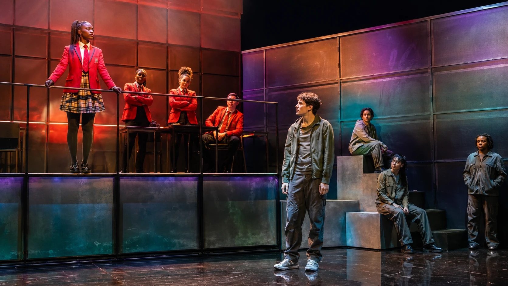 Sephy, a Black Cross student, elevated in a red school uniform while Callum, a white Nought student, stands in the lower acting space of a divided live-theatre set.